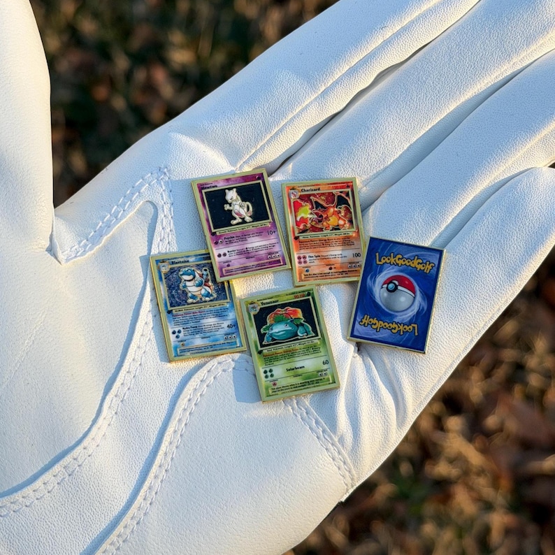 May include: A collection of gold-edged Pok&eacute;mon card pins, including Charizard, Blastoise, Venusaur, and Mewtwo, are displayed on a white golf glove. One pin features a Pok&eacute;ball design with the text "Look Good Golf."