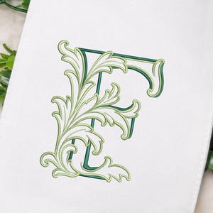 Puede incluir: Un artículo de tela blanca con la letra E bordada en un diseño decorativo de volutas de hojas. El bordado utiliza tonos de verde oscuro y claro con contornos finos, creando un monograma detallado de inspiración botánica en el tejido.