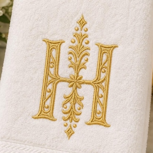 May include: A white cotton bath towel featuring a large, centred letter H embroidered in metallic gold thread. The letter is flanked by intricate, symmetrical floral and scrollwork patterns in the same gold colour, with a textured border at the bottom.