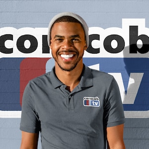 Può includere: Un uomo indossa una polo grigia con un logo che dice "corncob tv" in testo bianco su sfondo rosso e blu. Sta sorridendo e indossa un berretto di lana grigio.
