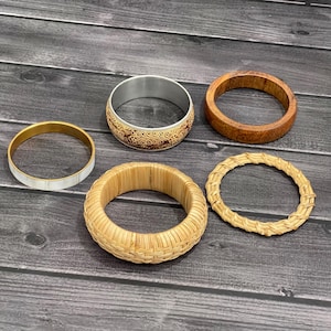 May include: A collection of five bangles including a thin white and gold-tone metal band, a wide metal band with an intricate brown floral pattern, a smooth dark brown wooden band, a thick woven rattan band, and a thin braided rattan band.