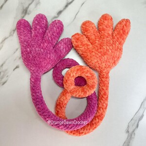 May include: Two plush, handmade crochet flowers in shades of pink and orange. The flowers have a textured, velvety appearance. The stems of the flowers are connected by a loop. The text "@OrangeDawnCrochet" is visible.