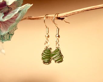 Pode incluir: Um par de brincos feitos à mão com vidro do mar verde envolto em arame prateado. Os brincos pendem de ganchos de orelha prateados. O fundo é um bege suave e quente, com uma flor e um galho desfocados no canto superior esquerdo.