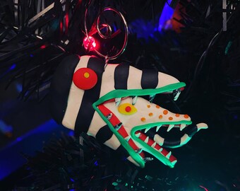 Puede incluir: Un adorno navideño hecho a mano con forma de criatura rayada de verde, rojo y blanco con una boca grande y dientes afilados. El adorno está hecho de arcilla polimérica y tiene un colgador de alambre.