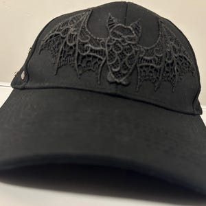 Può includere: Cappellino da baseball nero con un design a pipistrello dettagliato e simile al pizzo sul davanti. Il cappellino ha una visiera curva e piccole borchie argentate sul lato. Un accessorio alla moda.