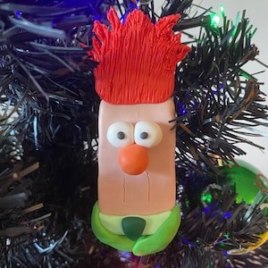 Peut inclure: Une décoration de Noël artisanale représentant un personnage de dessin animé avec des cheveux roux, une chemise verte et un visage rose. L'ornement est fait d'argile polymère et est suspendu à un arbre de Noël artificiel noir.