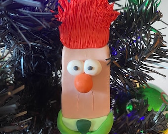 Puede incluir: Un adorno navideño hecho a mano con un personaje de dibujos animados con pelo rojo, una camisa verde y una cara rosa. El adorno está hecho de arcilla polimérica y está colgado de un árbol de Navidad artificial negro.