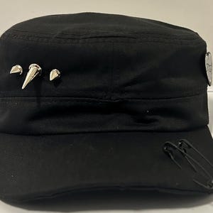 Może przedstawiać: Czarna czapka w stylu wojskowym ze srebrnymi metalowymi ćwiekami w kształcie stożków i ozdobnym emblematem. Czapka ma płaski daszek i jest ozdobiona dwiema czarnymi agrafkami. Stylowy dodatek.