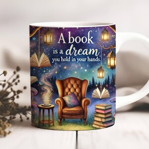 May include: White ceramic mug with a colorful design featuring a book, a cozy armchair, and the text "A book is a dream you hold in your hands."