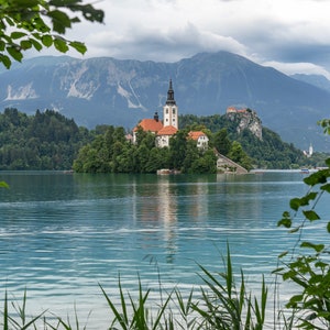 Può includere: Una vista pittoresca di una piccola isola con una chiesa sul lago di Bled in Slovenia. L'isola è circondata da lussureggianti alberi verdi e il lago è di un blu vibrante. Le montagne sullo sfondo sono coperte di nuvole.
