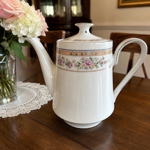 May include: A white porcelain teapot featuring a delicate floral band with pink and yellow flowers, green leaves, and soft peach-colored trim. The teapot has a curved spout, a matching lid with a small steam hole, and a silver-toned rim accent.