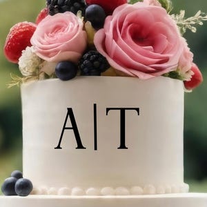 May include: A three-tiered white wedding cake decorated with fresh berries, pink roses, and greenery. The top tier features the letters "A|T" in black. Strawberries, blackberries, and blueberries adorn the cake, creating a festive and elegant design.