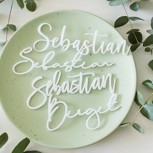 Op de afbeelding: Vier witte acryl placecards met de naam "Sebastian" in cursief schrift op drie van de kaarten en de naam "Drigit" in cursief schrift op de vierde kaart. De kaarten zijn gerangschikt op een lichtgroen bord met een witte achtergrond.