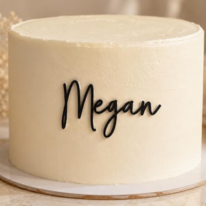 May include: A round cake with smooth, off-white frosting featuring a black script-style cake topper that reads Megan. The cake is positioned on a circular white base, showcasing a clean, minimalist presentation of the personalized name decoration.