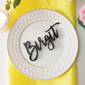 Op de afbeelding: Een wit bord met een gouden rand en een delicaat reli&euml;fpatroon. Op het bord ligt een zwart papieren plaatskaartje met de naam "Birgit" in cursief.