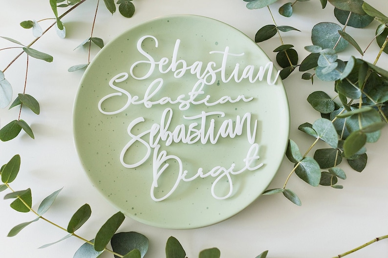 Op de afbeelding: Vier witte acryl placecards met de naam "Sebastian" in cursief schrift op drie van de kaarten en de naam "Drigit" in cursief schrift op de vierde kaart. De kaarten zijn gerangschikt op een lichtgroen bord met een witte achtergrond.