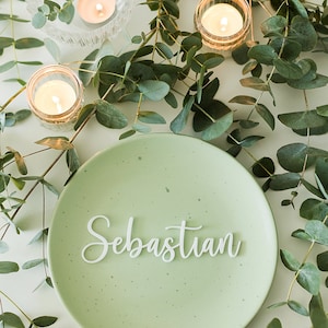 Op de afbeelding: Een groen bord met de naam Sebastian in witte, cursieve letters. Het bord is omringd door eucalyptusbladeren en kleine kaarsen.