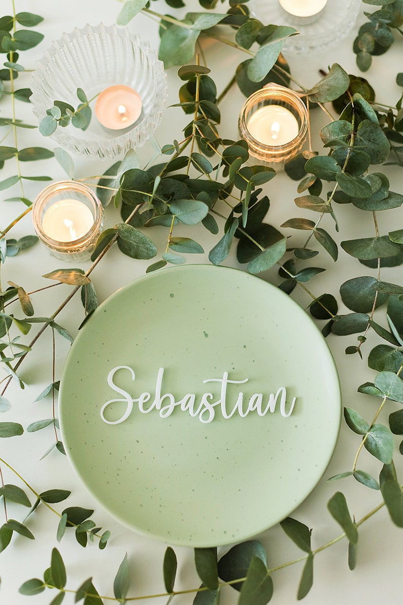 Op de afbeelding: Een groen bord met de naam Sebastian in witte, cursieve letters. Het bord is omringd door eucalyptusbladeren en kleine kaarsen.