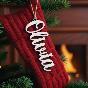 May include: A red knitted Christmas stocking with a wooden ornament that says "Olivia" hanging from a Christmas tree. The ornament is white and the name is written in a cursive font. The background includes a fireplace and Christmas tree branches.
