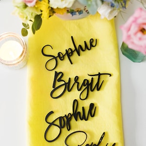 Może przedstawiać: Cztery czarne drewniane karty do miejsc z imionami Sophie, Birgit, Sophie i Sophie napisanymi kursywą na ż&oacute;łtym obrusie lnianym.