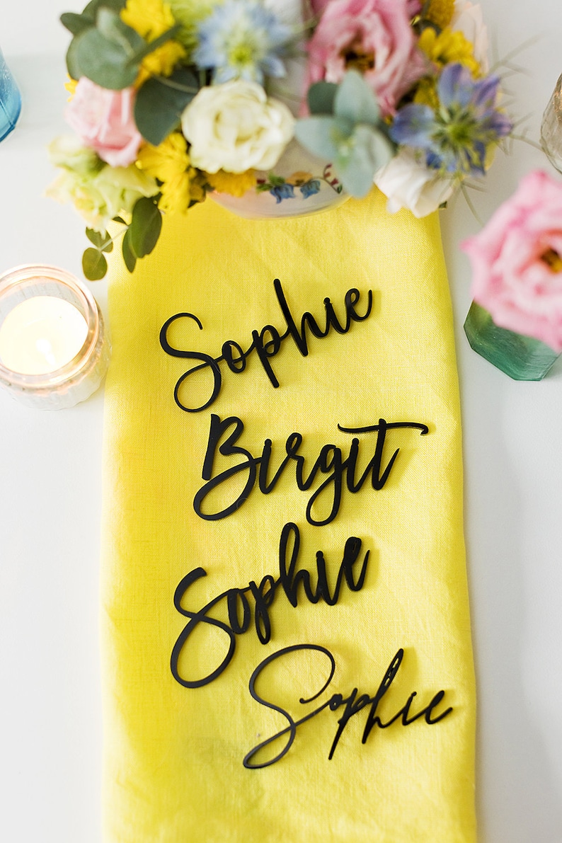 Może przedstawiać: Cztery czarne drewniane karty do miejsc z imionami Sophie, Birgit, Sophie i Sophie napisanymi kursywą na ż&oacute;łtym obrusie lnianym.