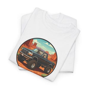 May include: A white t-shirt with a black and white illustration of a pickup truck in a desert landscape. The truck is in a circle with a red, orange, and blue background.