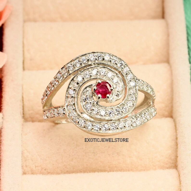 May include: A silver ring with a spiral design set with small white stones and a single red stone in the center. The ring is in a jewelry box.