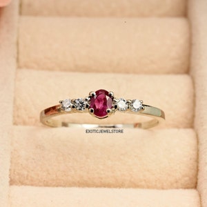May include: A silver ring featuring a red oval gemstone and smaller white gemstones on either side. The ring is displayed in a ring box.