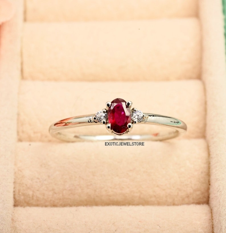 May include: A silver ring with a red oval ruby gemstone and two smaller white diamonds on either side. The ring is in a white box with a red velvet lining.