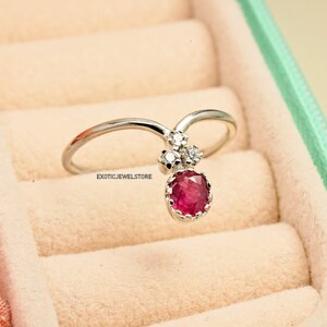 Pode incluir: Um anel de prata com uma grande gema de rubi redonda vermelha e diamantes brancos redondos menores. O anel &eacute; engastado em forma de V e o rubi fica pendurado no centro do V.