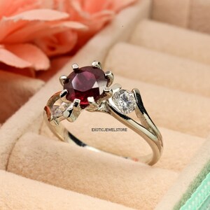 Op de afbeelding: Een zilveren ring met een rode edelsteen en twee kleinere witte edelstenen. De ring is gezet in een delicaat bloemmotief. "ExoticJewelStore" is gedrukt op de band.