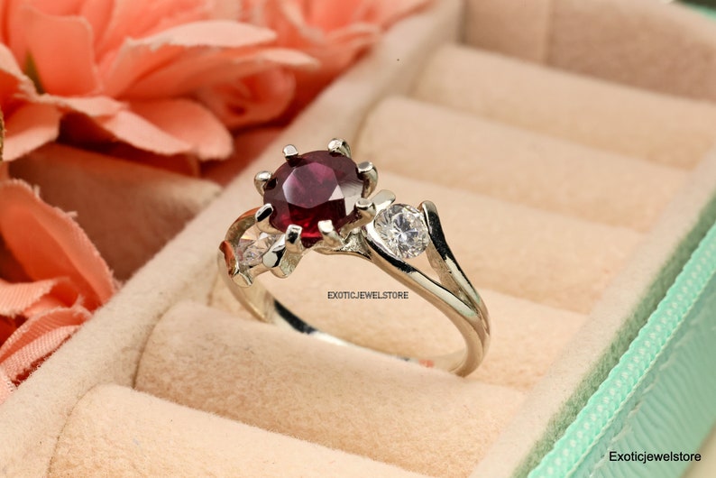 Op de afbeelding: Een zilveren ring met een rode edelsteen en twee kleinere witte edelstenen. De ring is gezet in een delicaat bloemmotief. "ExoticJewelStore" is gedrukt op de band.