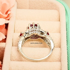 Op de afbeelding: Een zilveren ring met een cluster van rode edelstenen en kleine, heldere stenen. De ring heeft een gedetailleerd ontwerp met meerdere rode en heldere stenen. De ring is geplaatst tegen een zachte achtergrond.