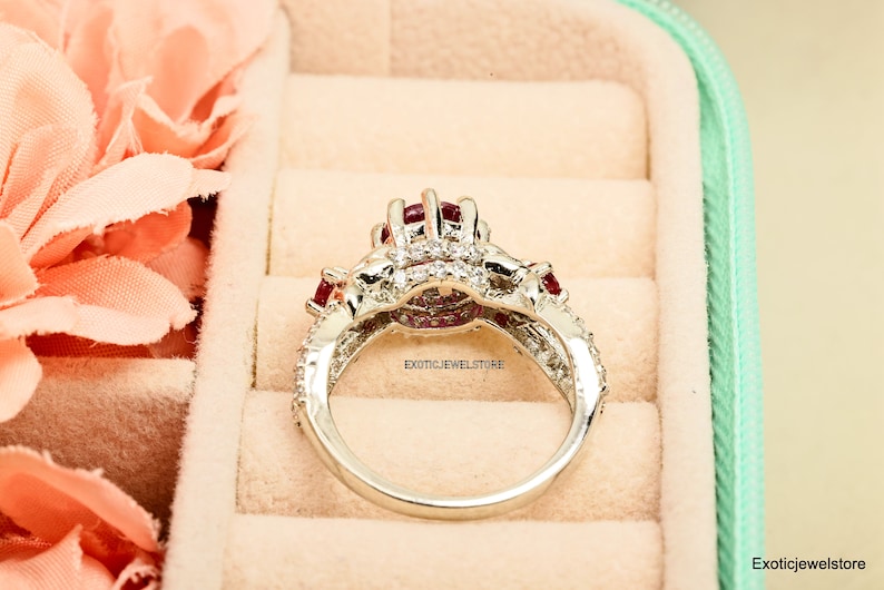 Op de afbeelding: Een zilveren ring met een cluster van rode edelstenen en kleine, heldere stenen. De ring heeft een gedetailleerd ontwerp met meerdere rode en heldere stenen. De ring is geplaatst tegen een zachte achtergrond.