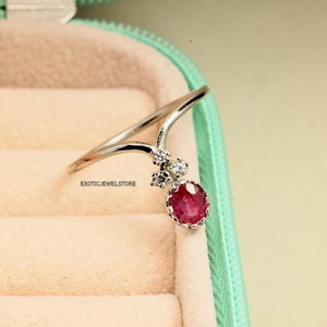 Pode incluir: Um anel de prata com uma gema vermelha e pequenas gemas transparentes. O anel est&aacute; em uma caixa de joias verde claro.