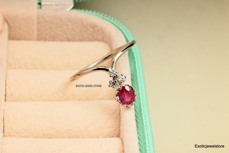 Pode incluir: Um anel de prata com uma gema vermelha e pequenas gemas transparentes. O anel est&aacute; em uma caixa de joias verde claro.