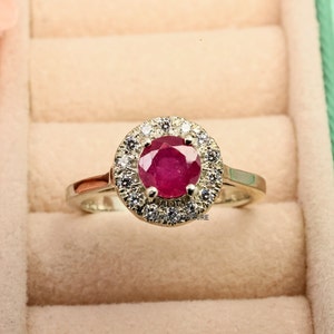 May include: A silver ring with a red ruby gemstone surrounded by a halo of smaller white diamonds.