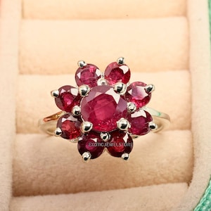 May include: A silver ring featuring a cluster of red rubies set in a flower design. The ring is displayed in a jewellery box.