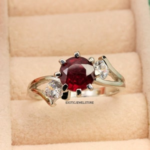 Op de afbeelding: Een zilveren ring met een grote rode edelsteen omringd door kleinere witte edelstenen. De ring heeft een delicaat, gebogen ontwerp.  "ExoticJewelStore" is op de ring gedrukt.