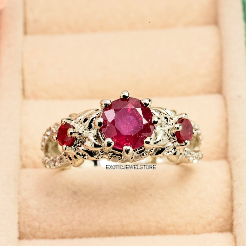 Op de afbeelding: Een zilveren ring met drie rode edelstenen. De centrale, ronde steen is groter, geflankeerd door twee kleinere stenen. De ring heeft een sierlijk, bloemig ontwerp met kleine, heldere accentstenen. De tekst "exoticjewelstore" is zichtbaar.