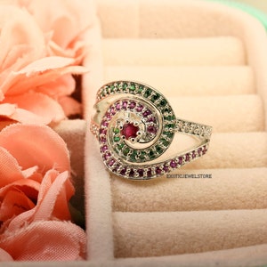 May include: A silver ring with a spiral design set with pink and green gemstones. The ring is in a white velvet ring box.