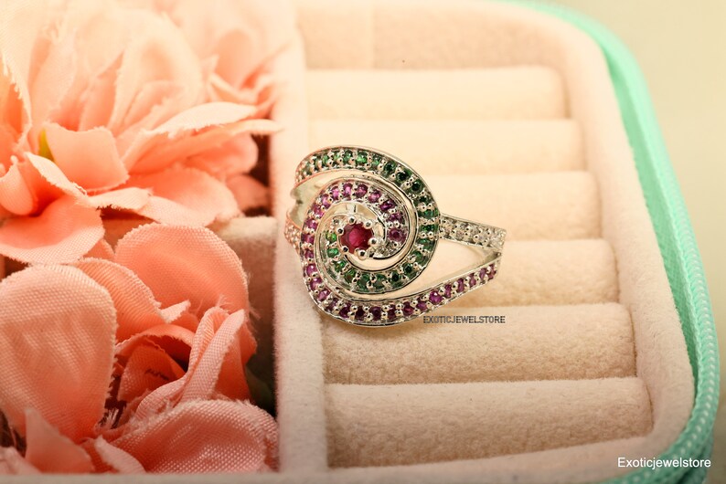 May include: A silver ring with a spiral design set with pink and green gemstones. The ring is in a white velvet ring box.