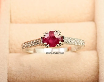 Puede incluir: Un anillo de plata con una piedra preciosa de rubí rojo y una banda de pequeños diamantes blancos. El anillo está en una caja de anillo.