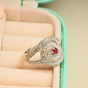 May include: A silver ring with a spiral design and a red gemstone in the center. The ring is set with clear cubic zirconia stones.