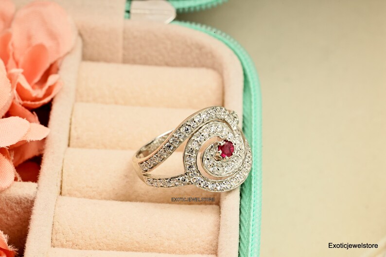 May include: A silver ring with a spiral design and a red gemstone in the center. The ring is set with clear cubic zirconia stones.