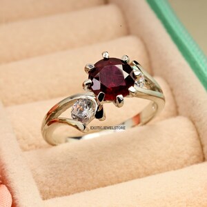 Op de afbeelding: Een zilveren ring met een grote, ronde, donkerrode edelsteen omringd door kleinere, heldere edelstenen. De ring is gezet op een delicate, gedraaide band.