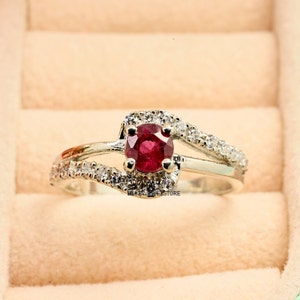 Peut inclure: Une bague en argent avec une pierre précieuse rouge entourée de petites pierres blanches. La bague a un design délicat et incurvé.