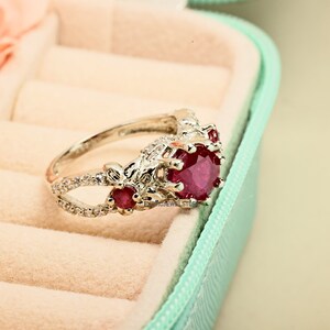 Op de afbeelding: Een zilveren ring met een grote, ronde, dieprode edelsteen. De ring heeft kleinere rode edelstenen en heldere accentstenen, met een bloemmotief op de band. De ring wordt gepresenteerd in een lichtblauwe sieradendoos.