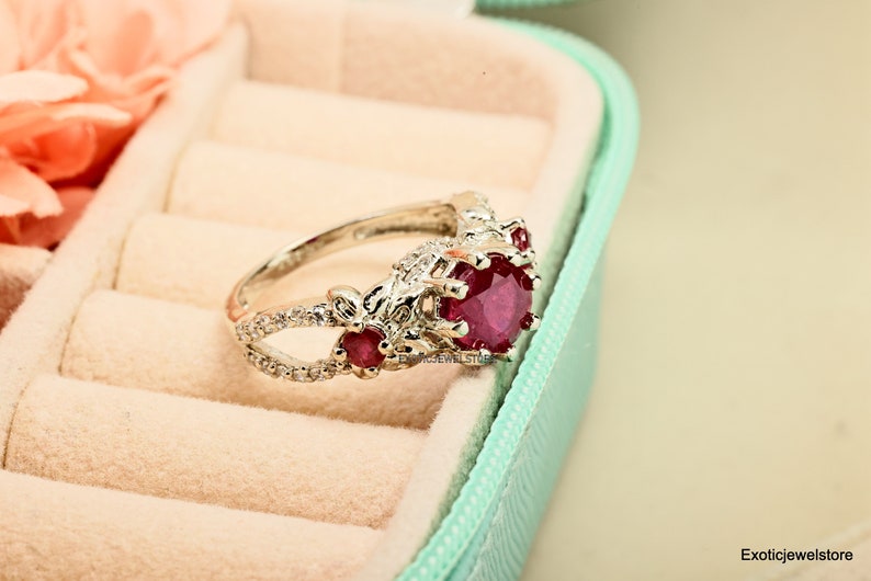 Op de afbeelding: Een zilveren ring met een grote, ronde, dieprode edelsteen. De ring heeft kleinere rode edelstenen en heldere accentstenen, met een bloemmotief op de band. De ring wordt gepresenteerd in een lichtblauwe sieradendoos.