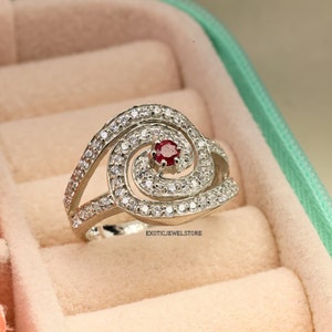 May include: A silver ring with a red gemstone in the center, surrounded by smaller white gemstones. The ring is set in a swirl design.